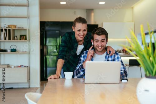 Obraz na płótnie A young married couple is talking to parents, family and friends on a video call via a laptop while sitting in the living room of their modern house. w salonie