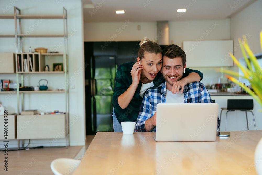 A young married couple is talking to parents, family and friends on a video call via a laptop while sitting in the living room of their modern house.