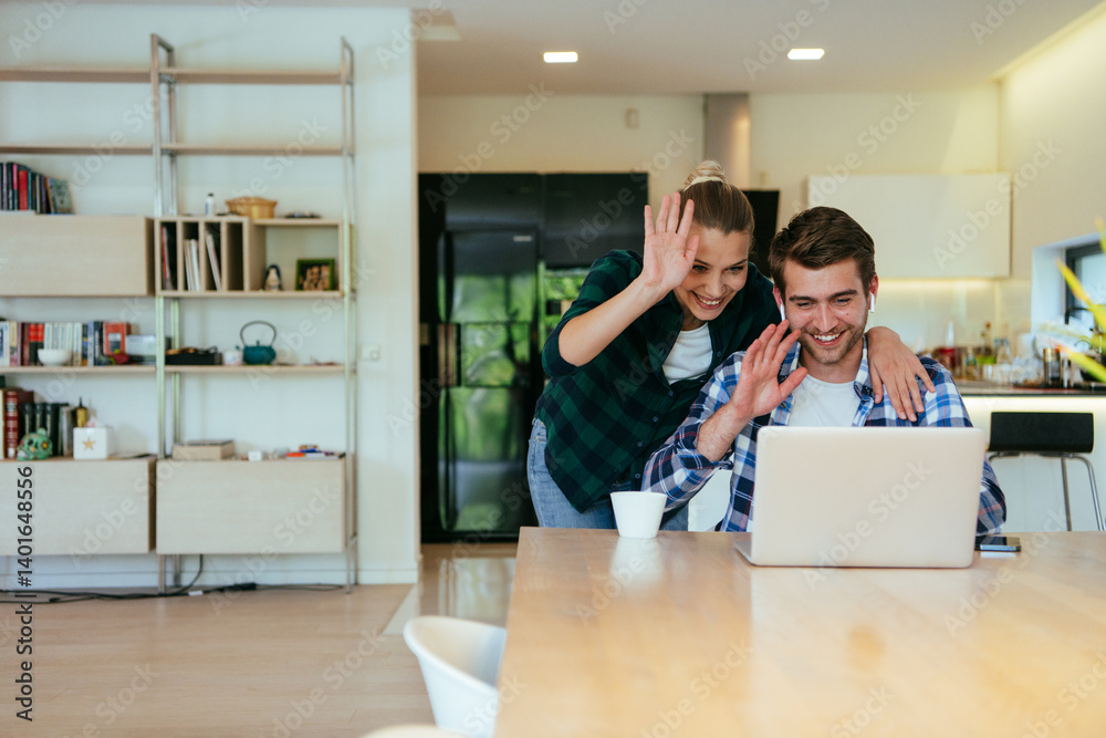 A young married couple is talking to parents, family and friends on a video call via a laptop while sitting in the living room of their modern house.