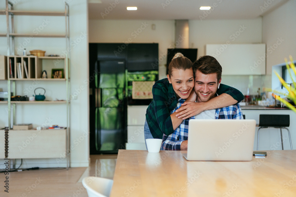 A young married couple is talking to parents, family and friends on a video call via a laptop while sitting in the living room of their modern house.