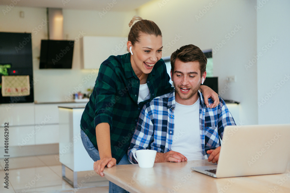 A young married couple is talking to parents, family and friends on a video call via a laptop while sitting in the living room of their modern house.