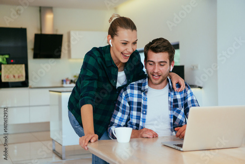Obraz na płótnie A young married couple is talking to parents, family and friends on a video call via a laptop while sitting in the living room of their modern house. w salonie