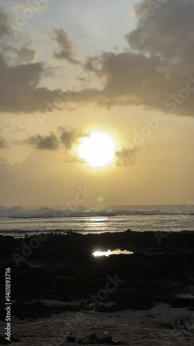 Golden sun illuminates a cloudy sky over the ocean waves. Dark volcanic rocks contrast with shimmering water in this serene coastal landscape.
