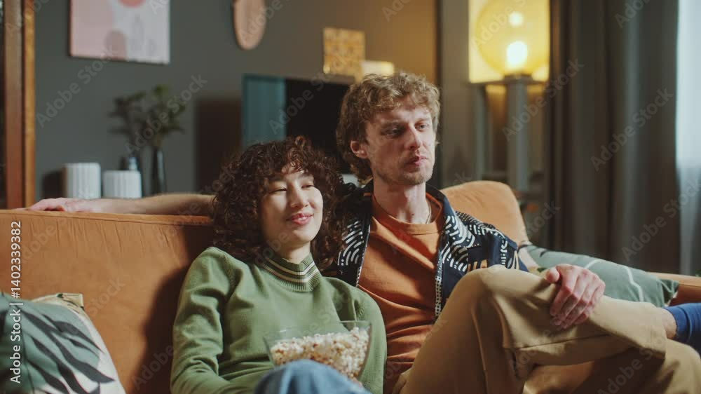 Young loving couple relaxing together on comfortable sofa, watching movie and sharing bowl of popcorn in cozy living room with warm lighting