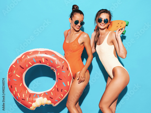 Two beautiful sexy smiling hipster women in summer swimwear bathing suits. Female in sunglasses. Positive models having fun with penny skateboards. With donut lilo inflatable mattress