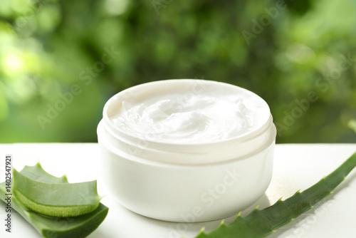 Moisturizing cream with extract of aloe vera in jar and cut leaves on white wooden table against blurred green background, closeup