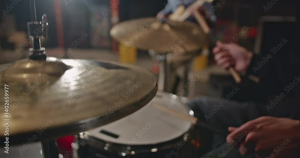 Close-up of a drummer playing the hi-hat and snare drum in a dimly lit room with a guitarist in the background.