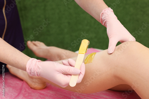 Professional cosmetologist applying hot depilatory wax on client's leg before hair removal procedure in beauty salon, closeup