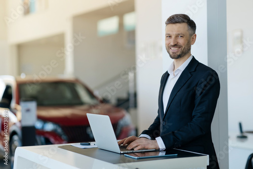Portrait of smiling Caucasian millennial car salesman working on laptop at car dealership showroom, handsome young man in suit standing at desk with computer and looking at camera, copy space