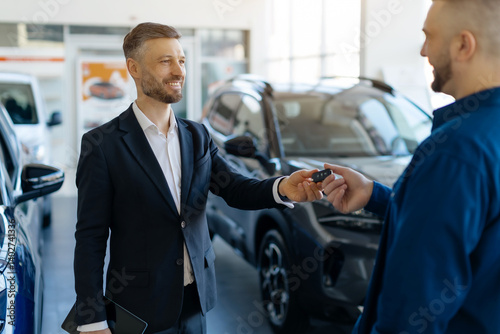 Smiling salesman giving car keys to male customer in bright modern car dealership showroom, auto salon manager in black suit greeting man with purchase, happy man buying vehicle