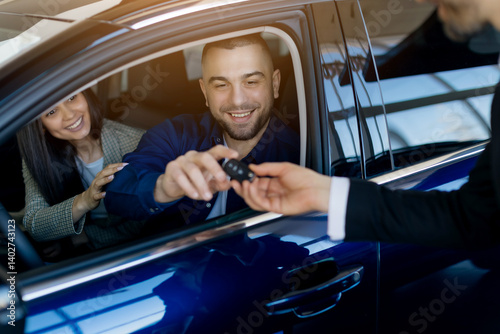 Smiling millennial couple receiving car keys from salesman while sitting inside their newly purchased car in dealership showroom, happy spouses buying modern vehicle in auto salon, closeup