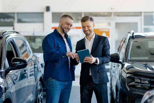 Caucasian millennial salesman showing digital tablet to a male customer, manager in suit and buyer standing between cars in modern dealership showroom, discussing auto characteristics