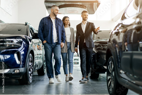 Smiling car salesman showing vehicles to a cheerful couple, buyers family and salon manager walking together inside spacious dealership showroom, considering options