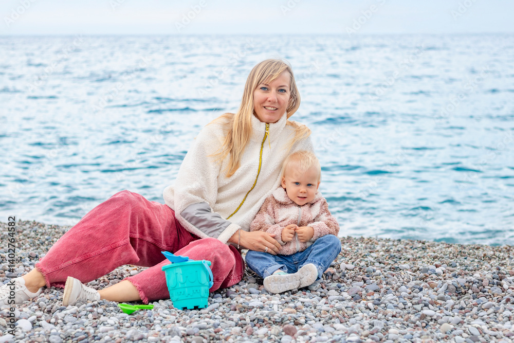 A joyful mother and her child play on the beach, surrounded by pebbles with the sea in the background.