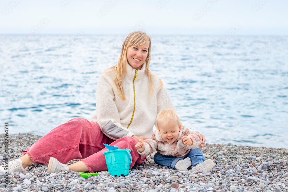 A joyful mother and her child play on the beach, surrounded by pebbles with the sea in the background.