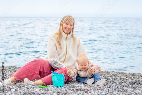 Obraz na płótnie A joyful mother and her child play on the beach, surrounded by pebbles with the sea in the background. w salonie