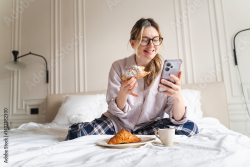 Young attractive cheerful woman in pajamas having breakfast with croissant and coffee in bed with mobile phone in hands