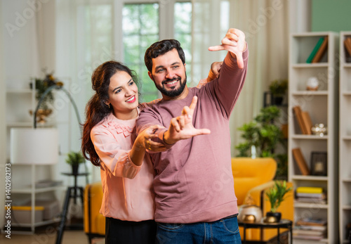 фотографія Happy Indian couple imagining house makeover with hand frame in cozy living room