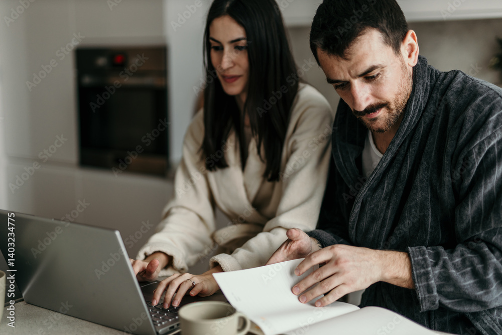 Young couple wearing bathrobes working from home using laptop and reading documents in the kitchen Young couple wearing bathrobes working from home using laptop and reading documents in the kitchen