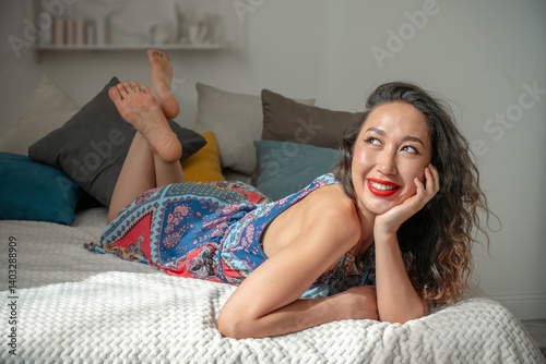 Young Asian woman in a light dress lies on her stomach on a bed in the rays of natural light and smiles