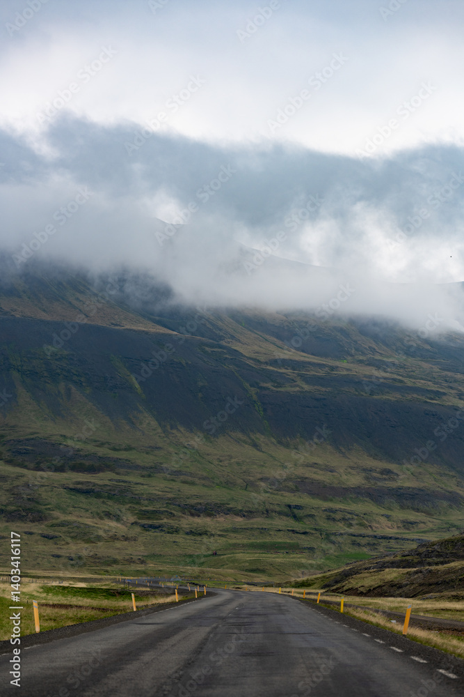 A winding road leads into misty Icelandic mountains, where low-hanging clouds brush rugged green slopes. The scene is tranquil and remote, evoking a sense of solitude and natural wonder.