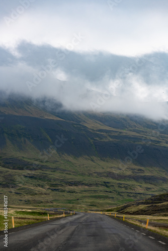 Obraz na płótnie A winding road leads into misty Icelandic mountains, where low-hanging clouds brush rugged green slopes. The scene is tranquil and remote, evoking a sense of solitude and natural wonder. w salonie