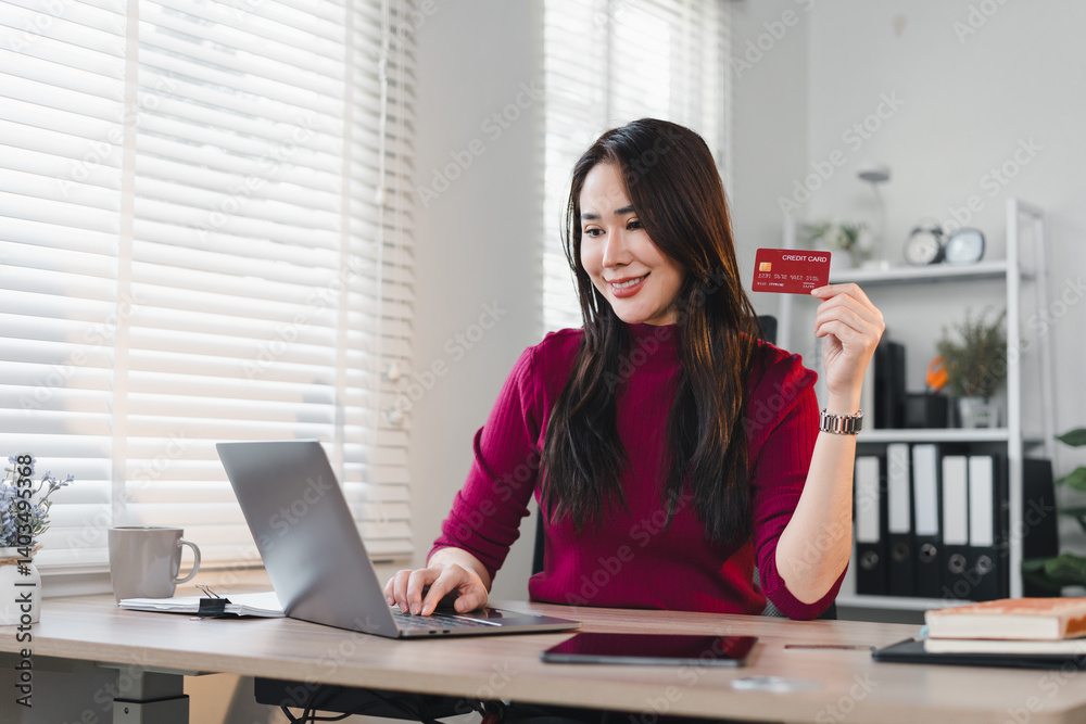 Asian businesswoman holding credit card while working on laptop in bright office, expressing confidence and focus Asian businesswoman holding credit card while working on laptop in bright office, expressing confidence and focus