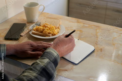 Man writing notes in notebook, eating scrambled eggs on toast, coffee beside smartphone at kitchen counter during morning routine