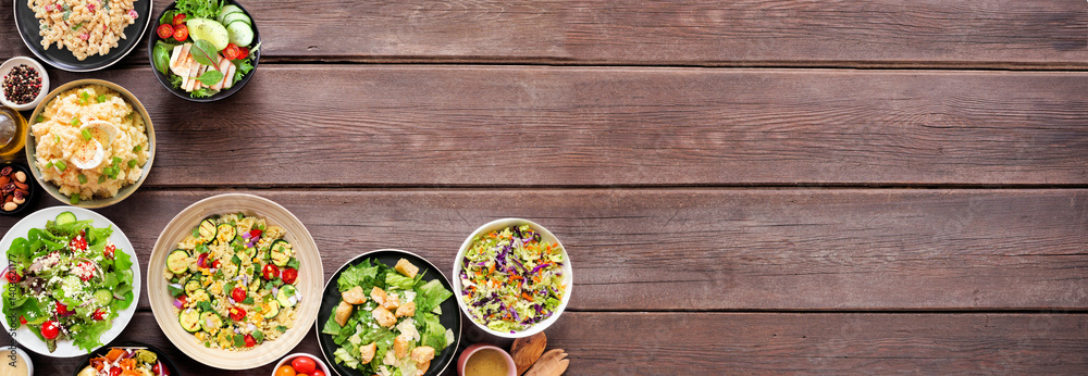 Assortment of delicious homemade salads. Overhead view corner border on a dark wood banner background. Summer food concept. Potato, caesar, pasta and green salads.