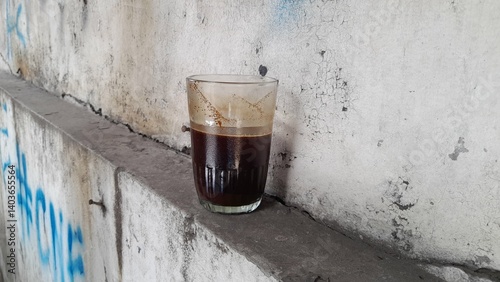 A small glass containing dark black coffee, placed on a narrow ledge against a rough, weathered wall with peeling paint. The coffee's dark color contrasts with the wall's texture