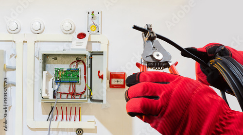 Electrician in protective gloves installs or repairs a fire alarm control system, preparing wires for connection, related to electrical and safety systems work.