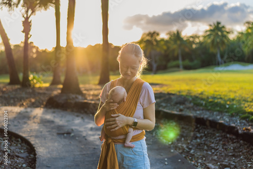 A loving mother walks through a peaceful park, carrying her baby securely in a sling. A heartwarming moment of maternal bonding, babywearing, and the joy of motherhood in nature