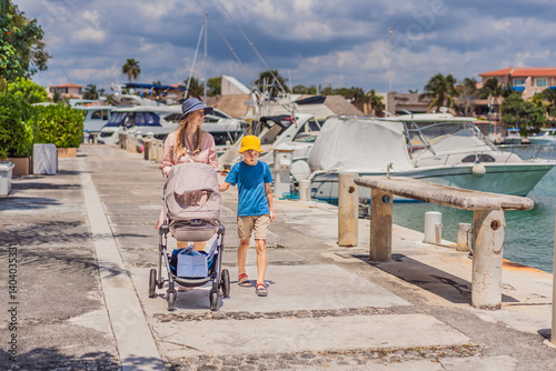 Obraz na płótnie A mother and her elder son enjoy a peaceful walk along the waterfront, pushing a baby stroller with their youngest family member. A heartwarming moment of family bonding, parenting, and outdoor w salonie