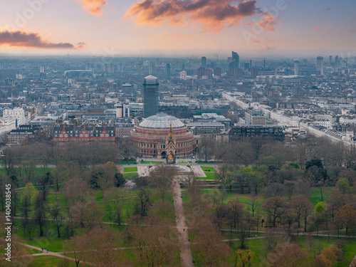 Aerial view of London's Royal Albert Hall with its domed roof, the Albert Memorial, surrounding parkland, and urban architecture under a cloudy sky.