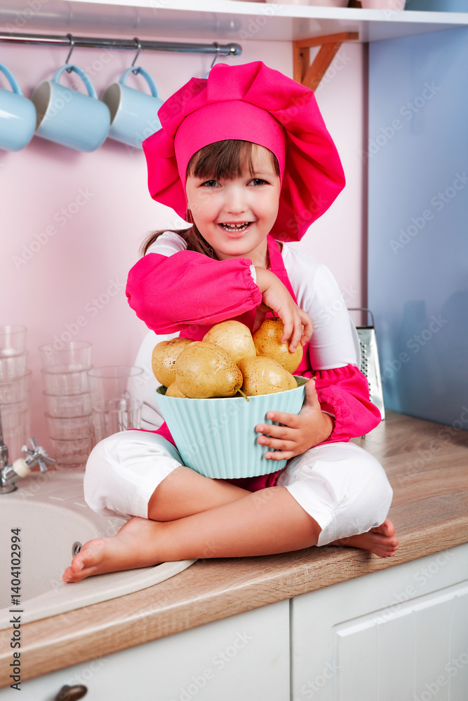 Happy little beautiful girl in chef costume holding bowl with ripe organic pears in hands while sitting in kitchen. Healthy eating and diet concept. Happy little beautiful girl in chef costume holding bowl with ripe organic pears in hands while sitting in kitchen. Healthy eating and diet concept.