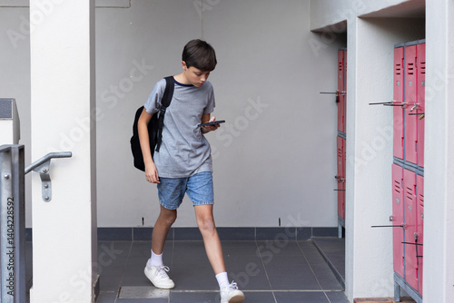 Walking in school hallway, boy with backpack using smartphone near lockers