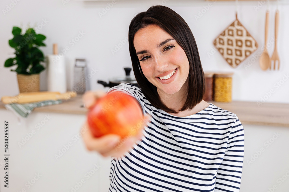 Young woman smiling and offering a fresh, ripe apple, promoting a healthy eating lifestyle in a modern kitchen setting Young woman smiling and offering a fresh, ripe apple, promoting a healthy eating lifestyle in a modern kitchen setting
