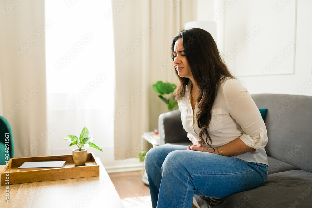 Sick latin woman experiencing a painful stomach ache while resting on the couch in the living room at home
