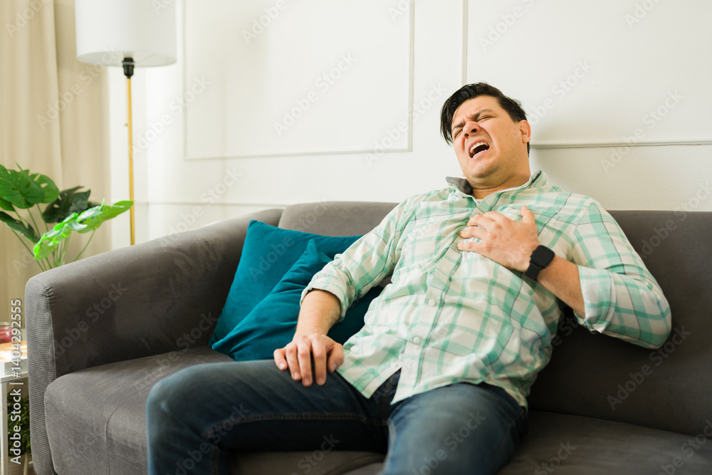 Hispanic man having a heart attack and touching his chest while experiencing painful angina and shortness of breath in his living room Hispanic man having a heart attack and touching his chest while experiencing painful angina and shortness of breath in his living room
