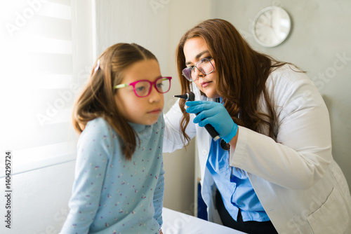 Female pediatrician wearing gloves using an otoscope examining ear of a little girl in doctor's office