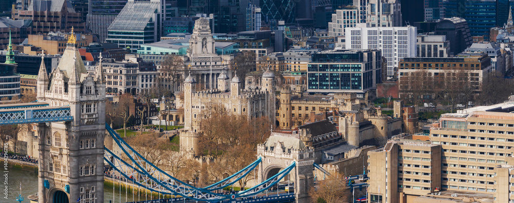 Obraz premium A detailed aerial view of Tower Bridge with its blue suspension elements, the historic Tower of London, and surrounding modern skyscrapers and buildings.
