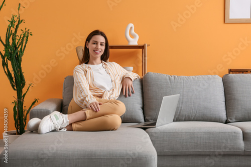 Smiling young woman relaxing on sofa with laptop, remote work and home comfor...