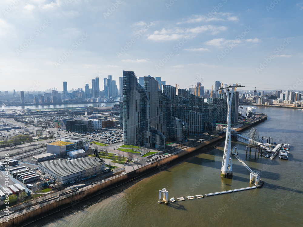 Aerial view of the Emirates Air Line cable car tower over the River Thames, with modern riverside buildings and Canary Wharf skyscrapers in the background.