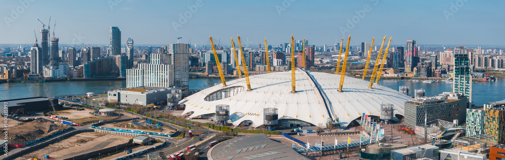 Aerial perspective of London highlighting the O2 Arena, modern buildings, construction sites, the River Thames, and Canary Wharf's high rises.