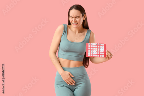 Sporty young woman with menstrual calendar suffering from cramps on pink background