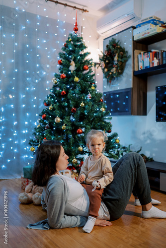 Little girl with pursed lips sits on her mom stomach lying on the floor near a decorated Christmas tree