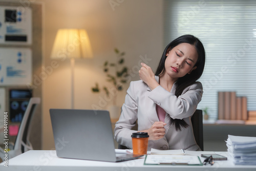 Young office worker massaging her shoulder, experiencing neck pain after long hours of working on a laptop at her workstation, feeling the strain of fatigue and stress