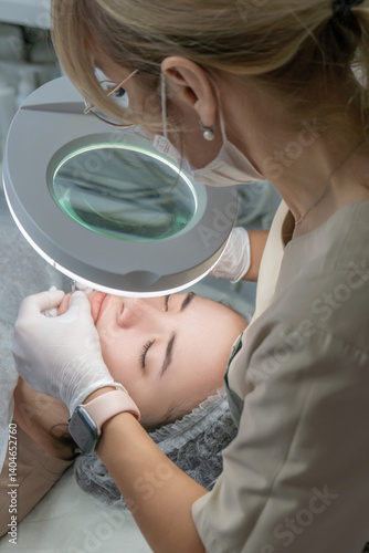 Cosmetology Clinic Face Treatment Procedure: Professional dermatologist performs facial treatment using magnifying lamp.