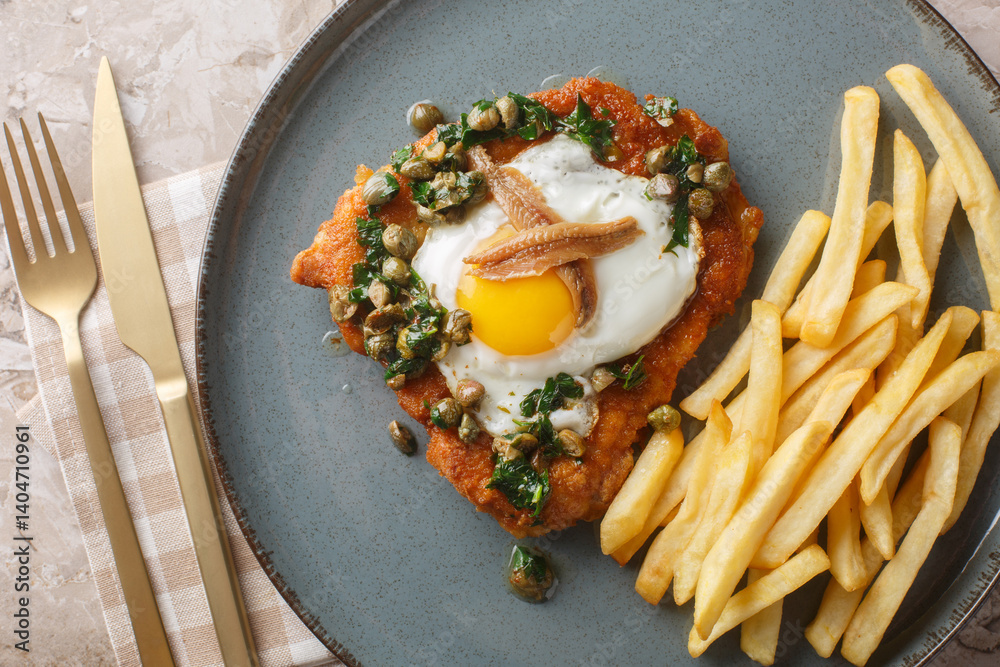 Schnitzel Holstein consists of a breaded cutlet served with a fried egg, anchovy and lemony caper sauce on top close up on the plate on the table. Horizontal top view from above