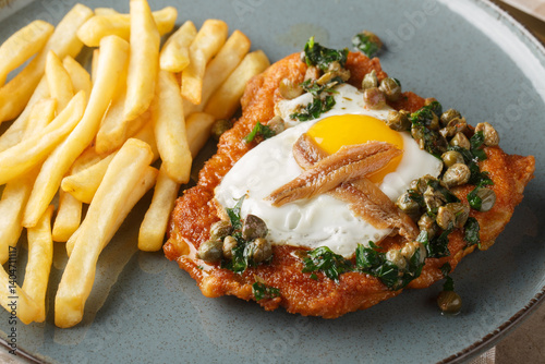 Obraz na płótnie Schnitzel Holstein consists of a breaded cutlet served with a fried egg, anchovy and lemony caper sauce on top close up on the plate on the table. Horizontal w salonie